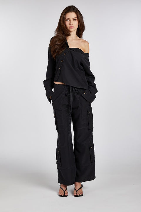 Modern and elegant one-shoulder asymmetric button-up blouse in black, crafted from buttery soft TENCEL modal woven fabric, shown as part of a matching set with baggy black cargo pants. Paired with heels and flip-flop heels, this sustainable, drapey top embodies minimal style and elevated essentials, offering a flattering fit with a relaxed yet subtly sexy silhouette. Designed for versatility, it resists wrinkles and transitions effortlessly from work to dinner, errands to events—perfect for any occasion.
