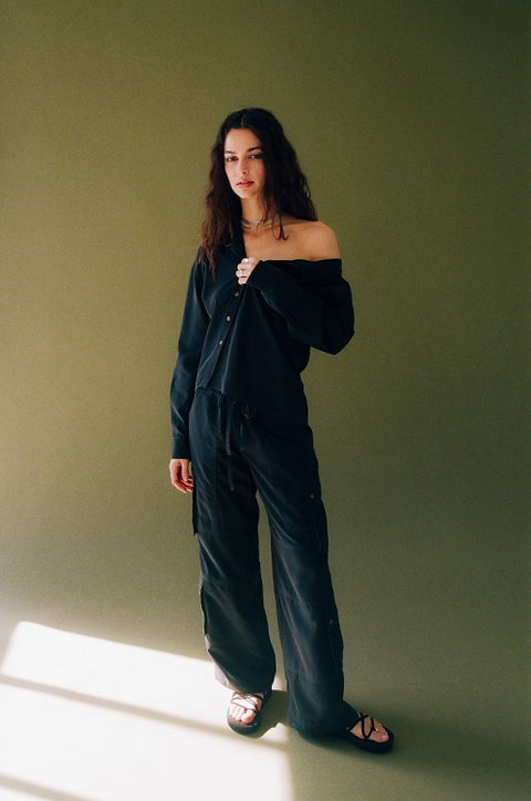 Modern and elegant one-shoulder asymmetric button-up blouse in black, crafted from buttery soft TENCEL modal woven fabric, shown as part of a matching set with baggy black cargo pants. Paired with heels and flip-flop heels, this sustainable, drapey top embodies minimal style and elevated essentials, offering a flattering fit with a relaxed yet subtly sexy silhouette. Designed for versatility, it resists wrinkles and transitions effortlessly from work to dinner, errands to events—perfect for any occasion.