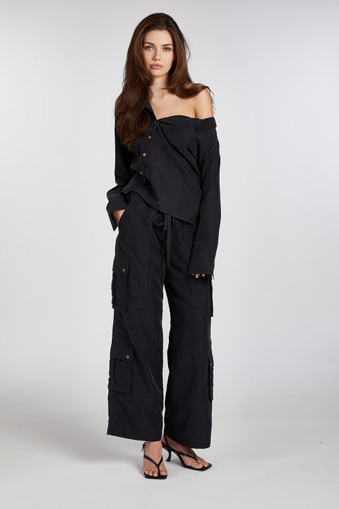 Model wearing a black one-shoulder asymmetric button-up blouse, paired with heels for an effortlessly chic look. Crafted from buttery soft, sustainable TENCEL modal fabric, this drapey and flattering blouse exudes modern elegance with a relaxed yet subtly sexy fit. Perfectly versatile for work, dinner, events, and beyond.