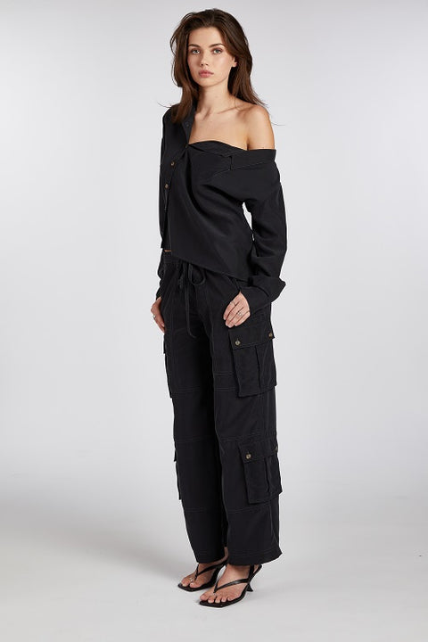 Angled shot of a model wearing a black one-shoulder asymmetric button-up blouse, exuding confidence and effortless allure. Crafted from buttery soft, sustainable TENCEL modal fabric, this drapey and flattering blouse highlights more skin for a subtly sexy yet elegant look. The perfect blend of modern sophistication and empowered femininity.