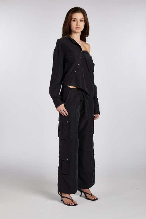 Angled view of modern and elegant one-shoulder asymmetric button-up blouse in black, crafted from buttery soft TENCEL modal woven fabric shown paired with a baggy black cargo pant and heels or stilettos . This sustainable, drapey top offers a flattering fit with a relaxed yet subtly sexy silhouette. Designed for versatility, it resists wrinkles and transitions effortlessly from work to dinner, errands to events—perfect for any occasion.