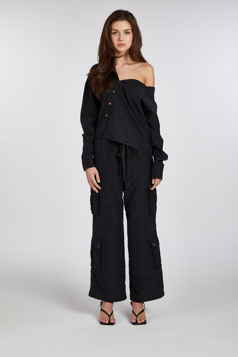 Modern and elegant one-shoulder asymmetric button-up blouse in black, crafted from buttery soft TENCEL modal woven fabric, shown as part of a matching set with baggy black cargo pants. Paired with heels and flip-flop heels, this sustainable, drapey top embodies minimal style and elevated essentials, offering a flattering fit with a relaxed yet subtly sexy silhouette. Designed for versatility, it resists wrinkles and transitions effortlessly from work to dinner, errands to events—perfect for any occasion.