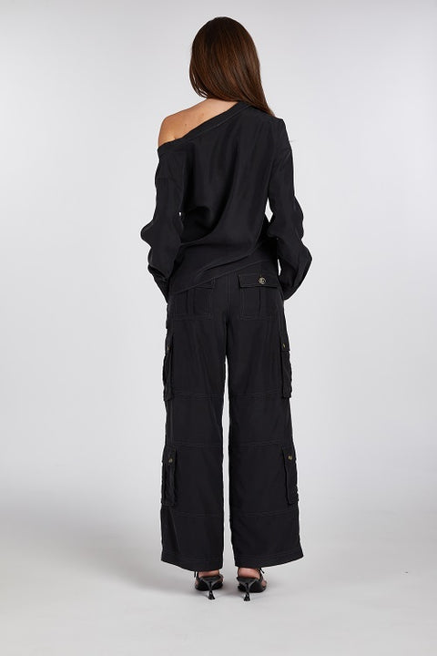 Back view of modern and elegant one-shoulder asymmetric button-up blouse in black, crafted from buttery soft TENCEL modal woven fabric, shown paired with heels and flip-flop heels. This sustainable, drapey top embodies minimal style and elevated essentials, offering a flattering fit with a relaxed yet subtly sexy silhouette. Designed for versatility, it resists wrinkles and transitions effortlessly from work to dinner, errands to events—perfect for any occasion.