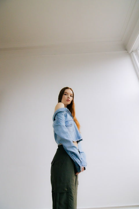 Side view of off-shoulder denim jacket with a built-in white ribbed racerback tank top, paired with drapey, baggy green cargo pants for an effortlessly cool look. Crafted from premium Italian denim in a light wash, this slouchy yet elegant 2-in-1 top features a slightly cropped fit, making it the perfect transitional outfit. Designed for versatility, it’s ideal for summer nights or layering through the seasons.