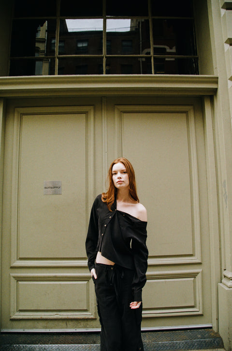 Modern and elegant one-shoulder asymmetric button-up blouse in black, crafted from buttery soft TENCEL modal woven fabric, shown as part of a matching set with baggy black cargo pants in SoHo, NYC. Paired with heels and flip-flop heels, this sustainable, drapey top embodies minimal style and elevated essentials, offering a flattering fit with a relaxed yet subtly sexy silhouette. Designed for versatility, it resists wrinkles and transitions effortlessly from work to dinner, errands to events—perfect for any