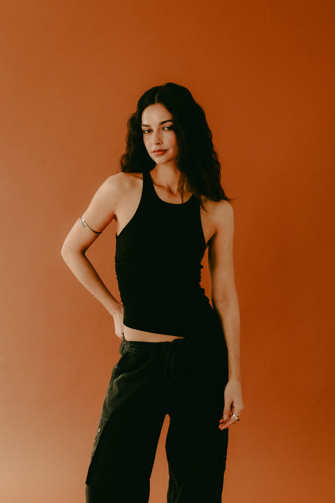 Black-on-black outfit featuring a chic and elegant black racerback tank top with an asymmetric hem, paired with baggy black cargo pants. Made from soft, stretchy, and sustainable ribbed knit fabric for a comfortable, flattering fit. Designed for versatility—it can be styled for a sexy, body-hugging look or worn more full coverage for effortless sophistication. Pair with heels, sneakers, sandals, or boots for any occasion.