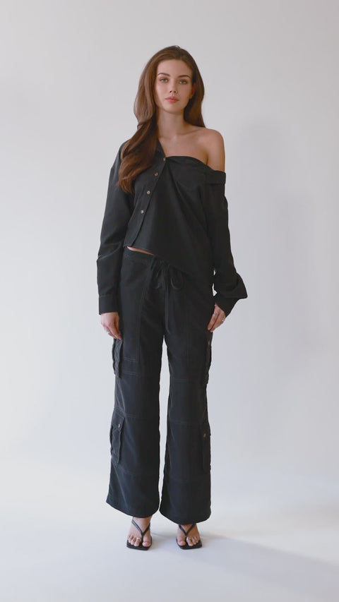 360-degree video of an elegant one-shoulder asymmetric button-up blouse in black, crafted from buttery soft TENCEL modal woven fabric, shown as part of a matching set with baggy black cargo pants in SoHo, NYC. Paired with heels and flip-flop heels, this sustainable, drapey top embodies minimal style and elevated essentials, offering a flattering fit with a relaxed yet subtly sexy silhouette. Designed for versatility, it resists wrinkles and transitions effortlessly from work to dinner, errands to ev